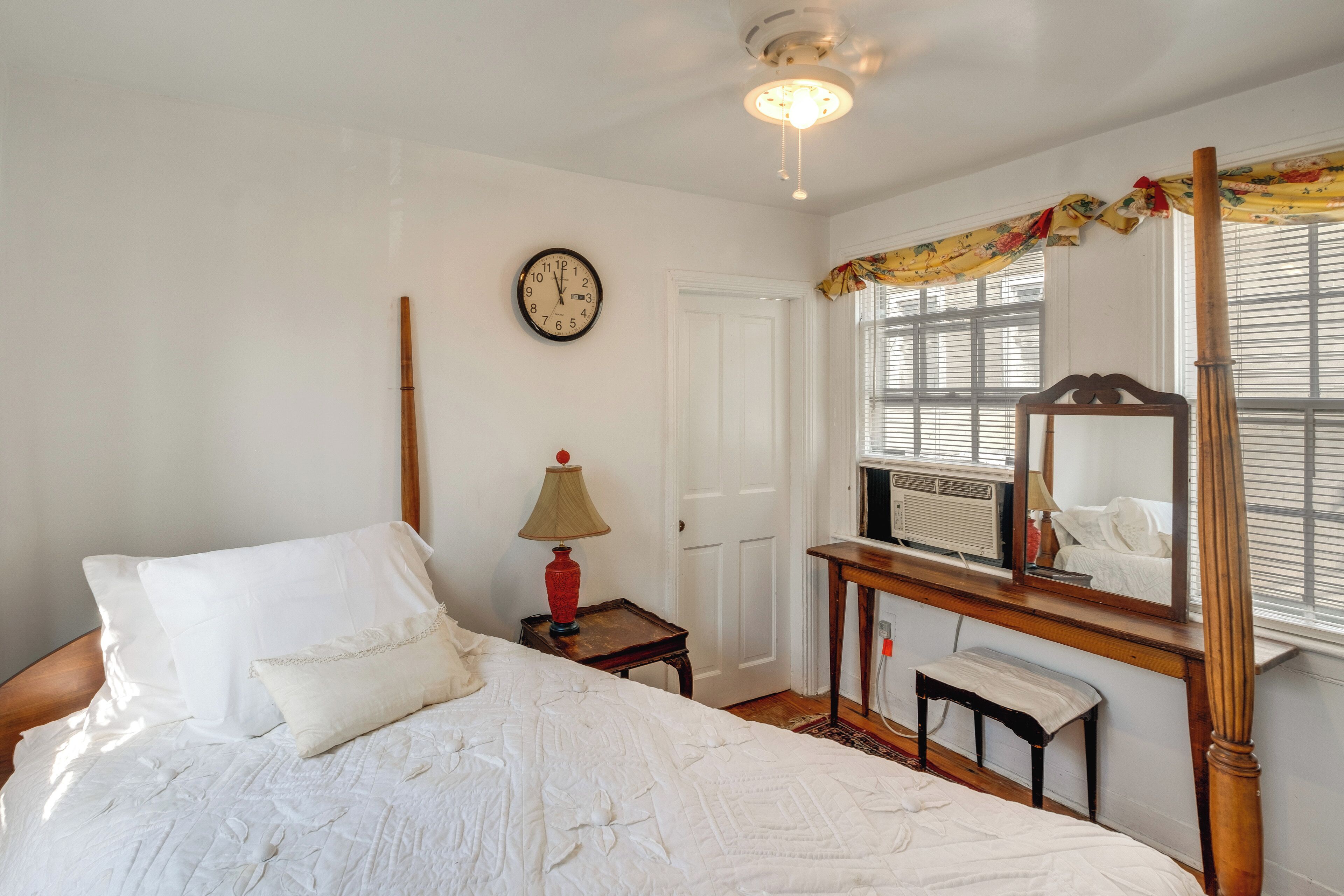 Une chambre à coucher avec un lit, une table de chevet avec une lampe, une horloge, un miroir et une fenêtre avec des stores.