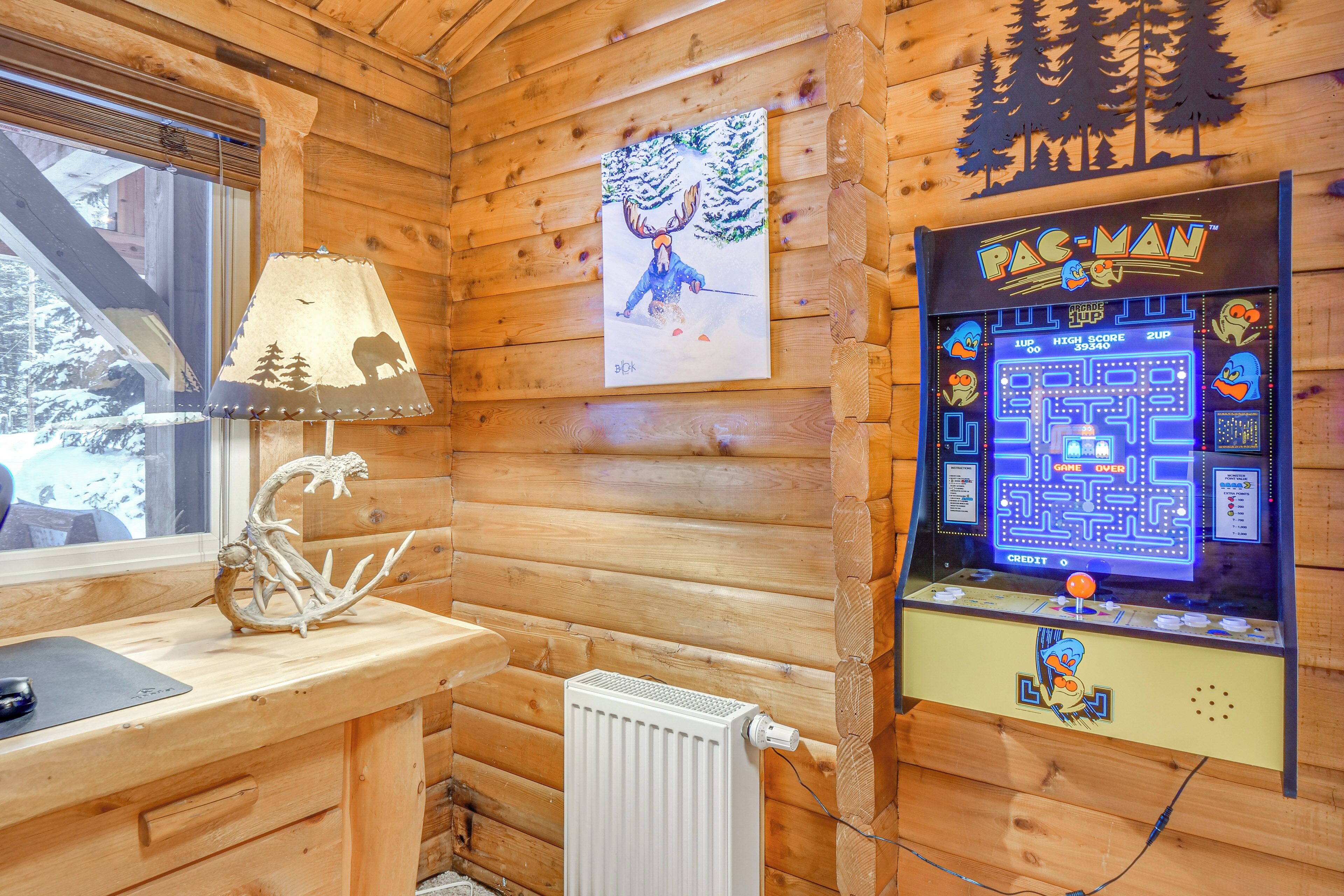A wooden cabin interior with a Pac-Man arcade game, a lamp, a deer head decoration, and a window with a view of snow-covered trees.