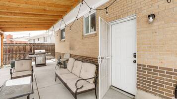 An outdoor patio with a brick wall, a white door, a sofa, a table, and a grill.