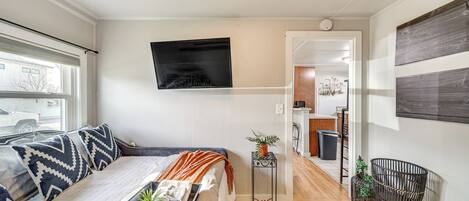 A modern bedroom with a bed, a TV, a small table with a plant, and a view of the kitchen.