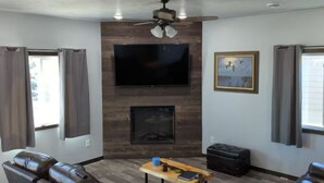 A living room with a fireplace, leather sofas, a wooden coffee table, and a flat-screen TV mounted on the wall. - Charming 3-bedroom house with WiFi, AC minutes from Lake Oahe/ 3 stall garage (Pierre)