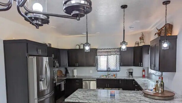 A modern kitchen with dark cabinetry, a stainless steel refrigerator, a granite countertop island with stools, pendant lighting, and a window with patterned curtains. - Charming 3-bedroom house with WiFi, AC minutes from Lake Oahe/ 3 stall garage (Pierre)