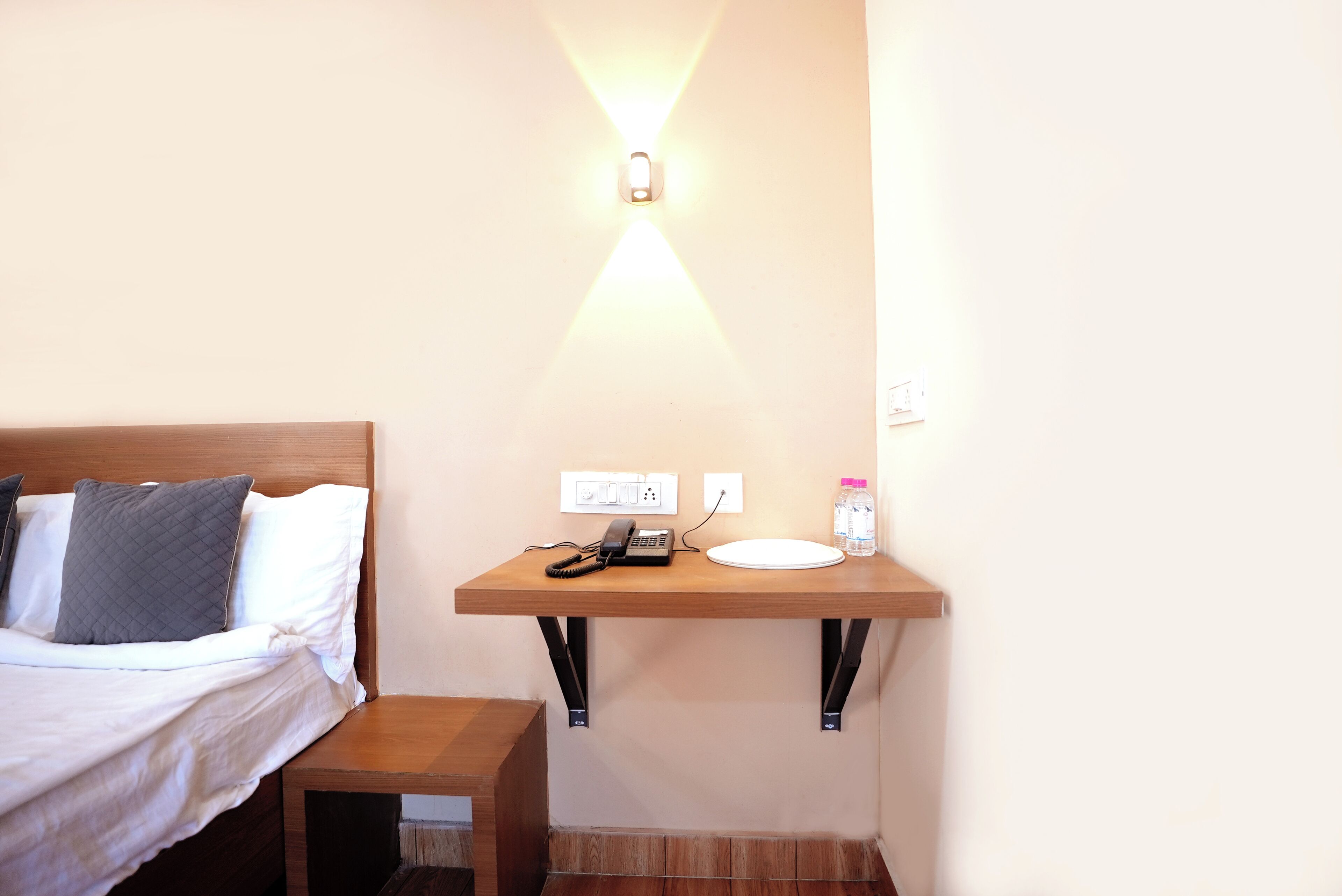 A hotel room with a bed, bedside table, and a small desk with a phone and a bottle.