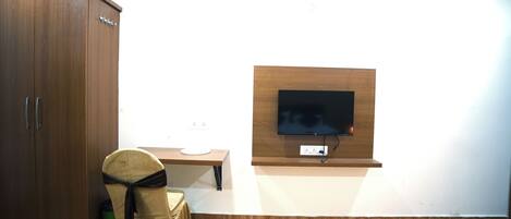 A room with a wooden wardrobe, a small table, a chair with a black cover, and a mounted TV on the wall.