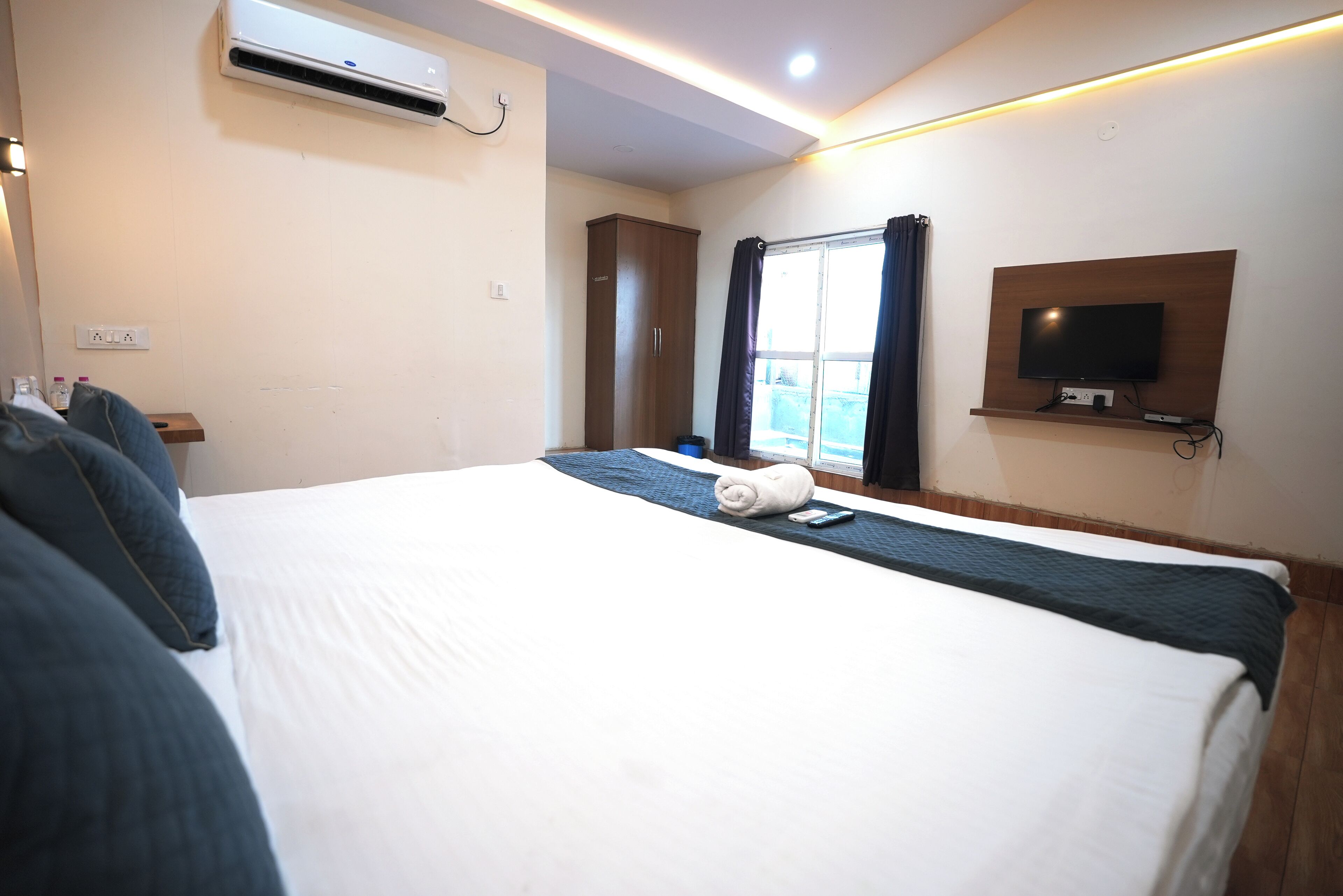 A hotel room with a large bed, a flat-screen TV mounted on the wall, an air conditioning unit above the bed, and a window with curtains.