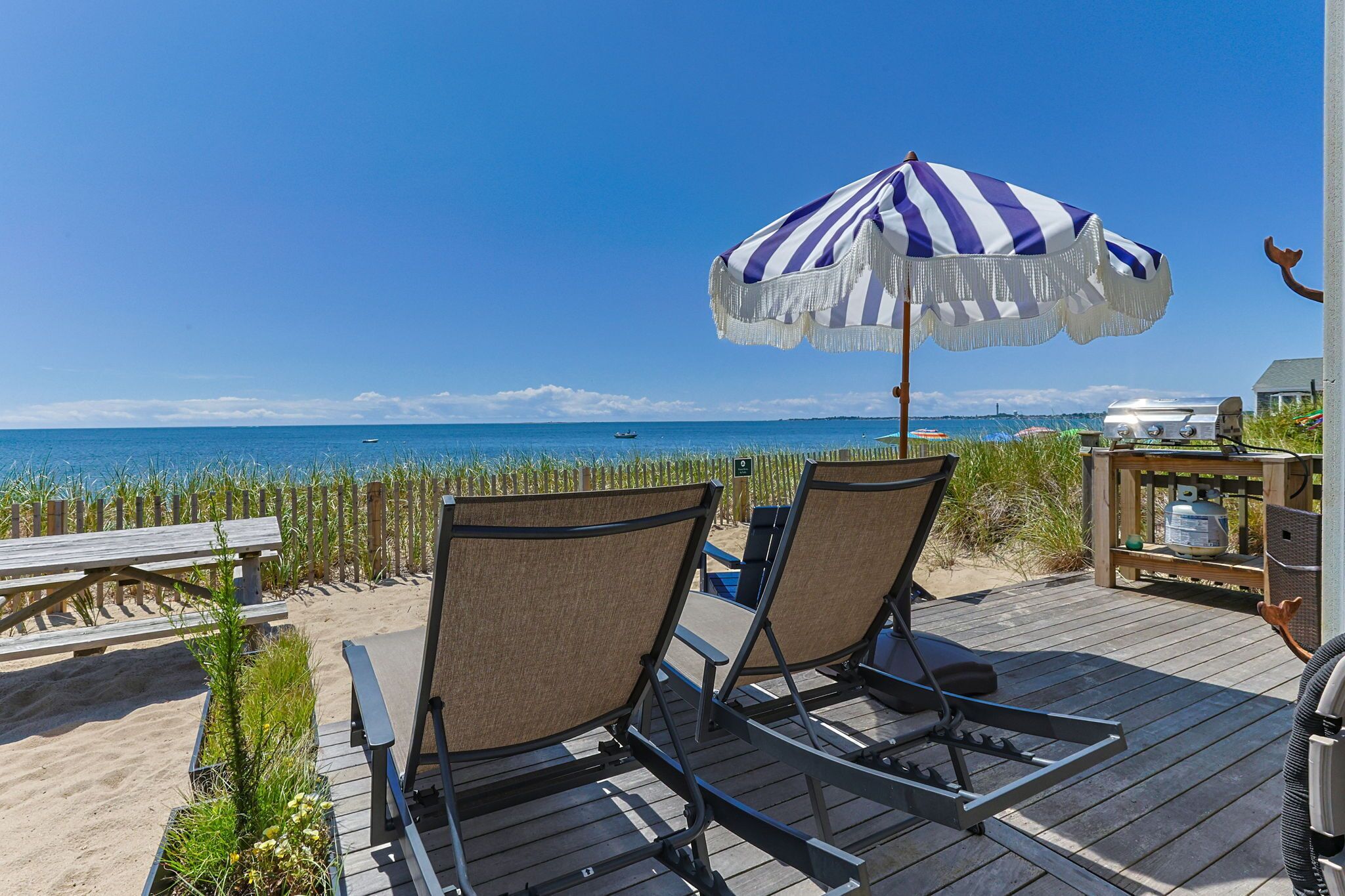 A deck with two lounge chairs and a striped umbrella overlooking a beach.