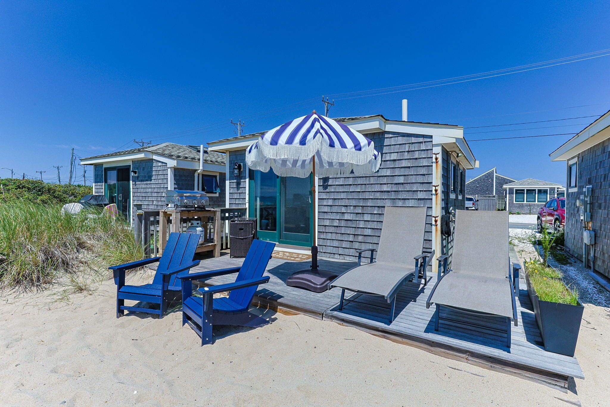 A beachfront property with a covered outdoor seating area, a barbecue grill, and a small building with a blue and white striped umbrella.