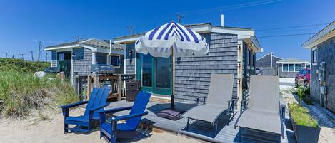A beachfront property with a covered outdoor seating area, a barbecue grill, and a small building with a blue and white striped umbrella.