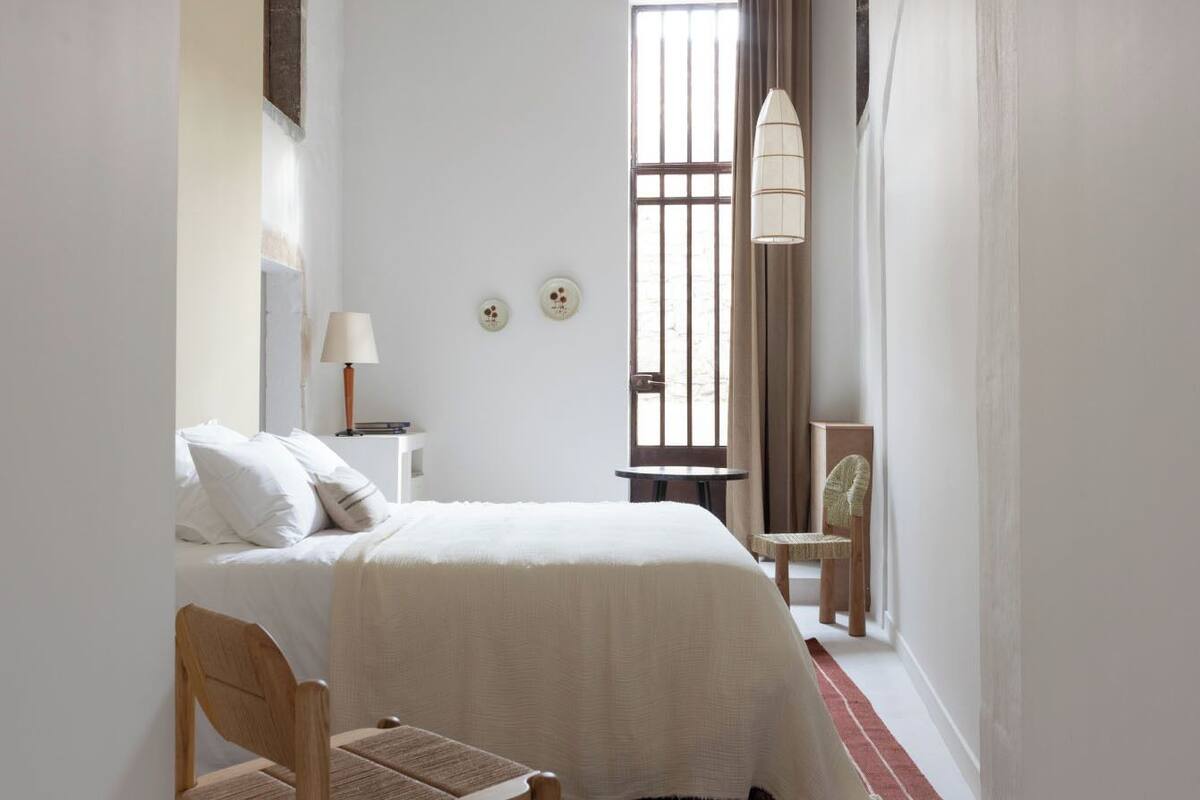 A bedroom with a bed, a chair, a nightstand, a lamp, and a window with curtains.