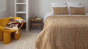 Une chambre avec un plafond en pente, un lit avec une literie blanche et marron, une chaise jaune avec un coussin à motifs, une petite table de chevet avec des livres et une lampe, et un luminaire suspendu.