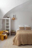 Un dormitorio con techo inclinado, cama con ropa de cama blanca y marrón, una silla amarilla con cojín estampado, una mesita de noche pequeña con libros y una lámpara, y una lámpara colgante.