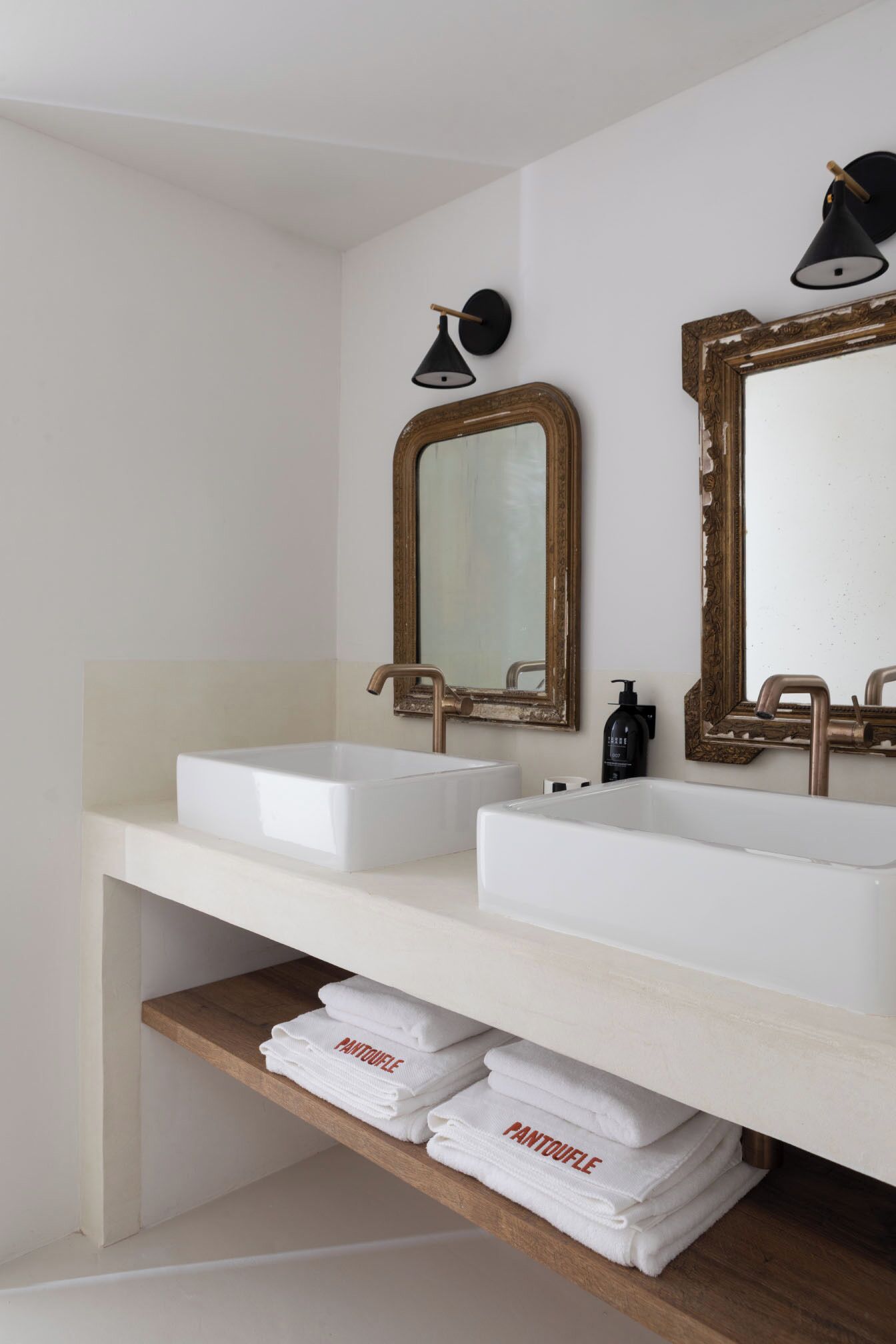 Une salle de bain avec deux vasques blanches, une étagère en bois où sont pliées des serviettes, et deux miroirs muraux dotés de cadres décoratifs.