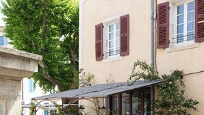 Un café en plein air avec des tables et des chaises, une petite fontaine et, au fond, un bâtiment aux volets rouges.
