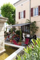 Un café al aire libre con mesas y sillas, una pequeña fuente de agua y un edificio con contraventanas rojas al fondo.
