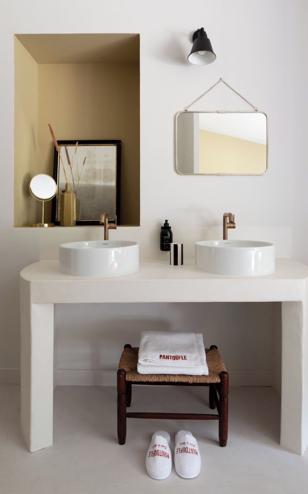 A bathroom with two sinks, a mirror, a small stool, and a pair of slippers.