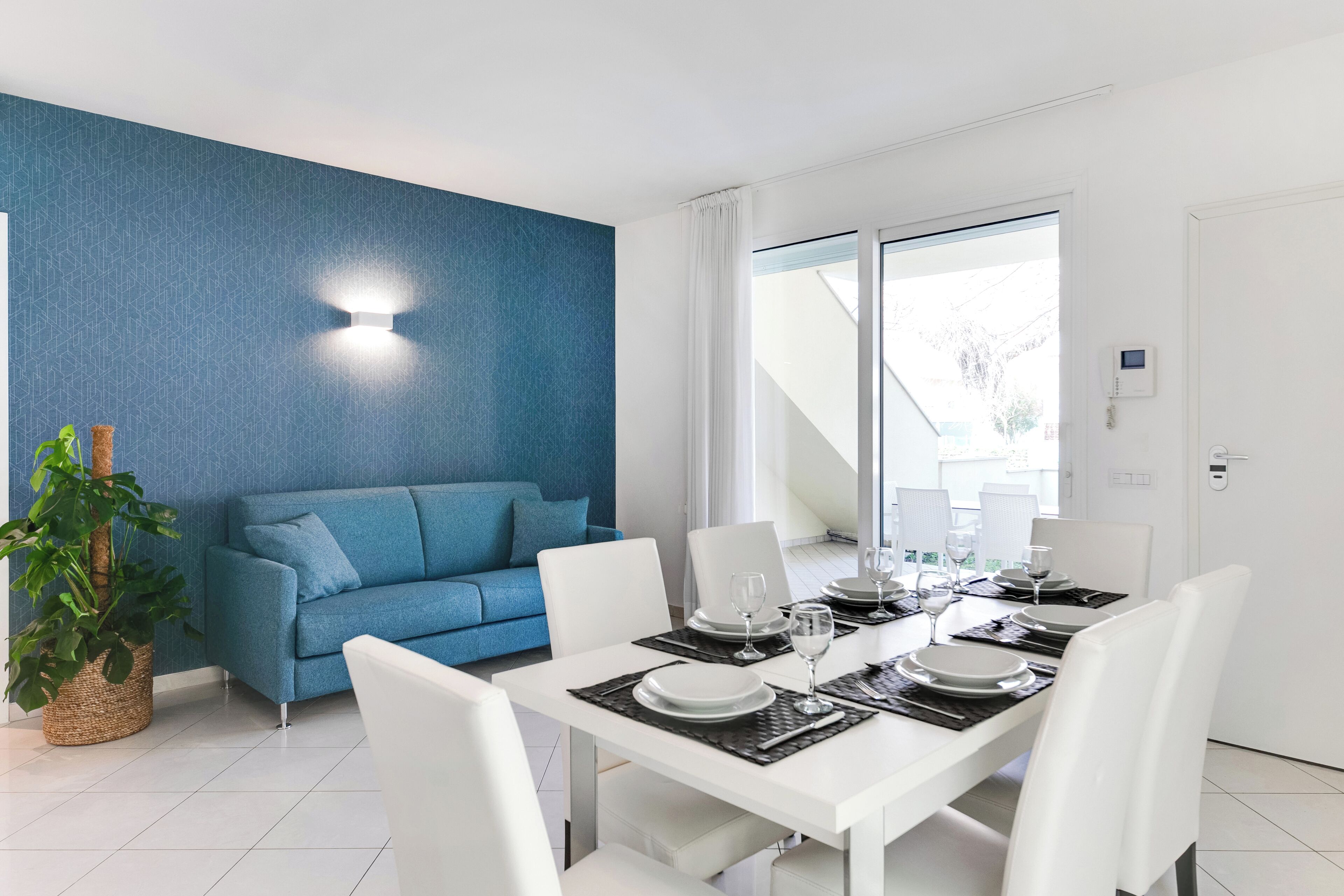 Ein moderner Essbereich mit einem weißen Tisch, gedeckt für vier Personen, einem blauen Sofa und einer Zimmerpflanze.