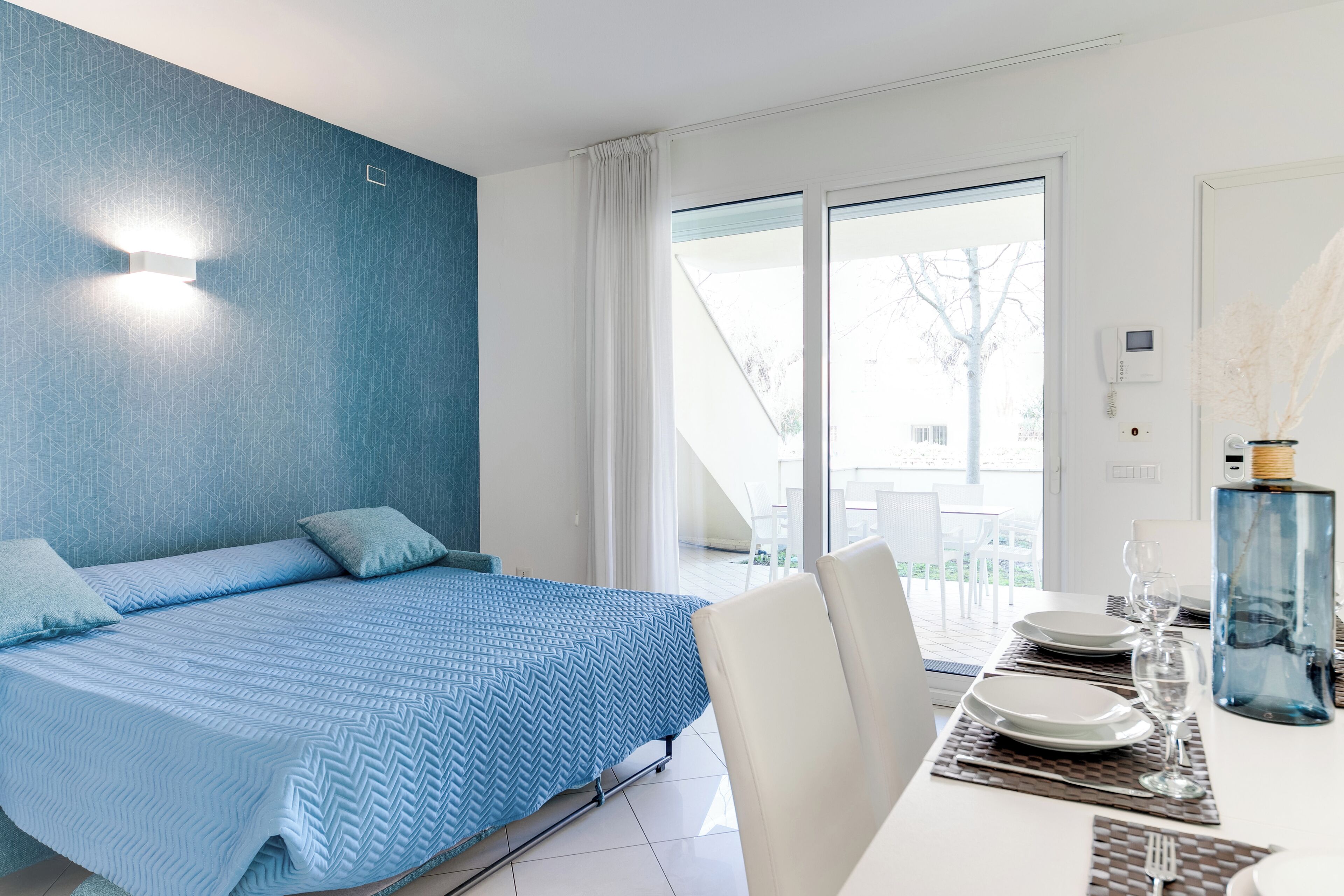 Habitación de hotel moderna con una cama grande, una zona de comedor con una mesa preparada para dos personas, y vistas al exterior a través de una puerta corredera.