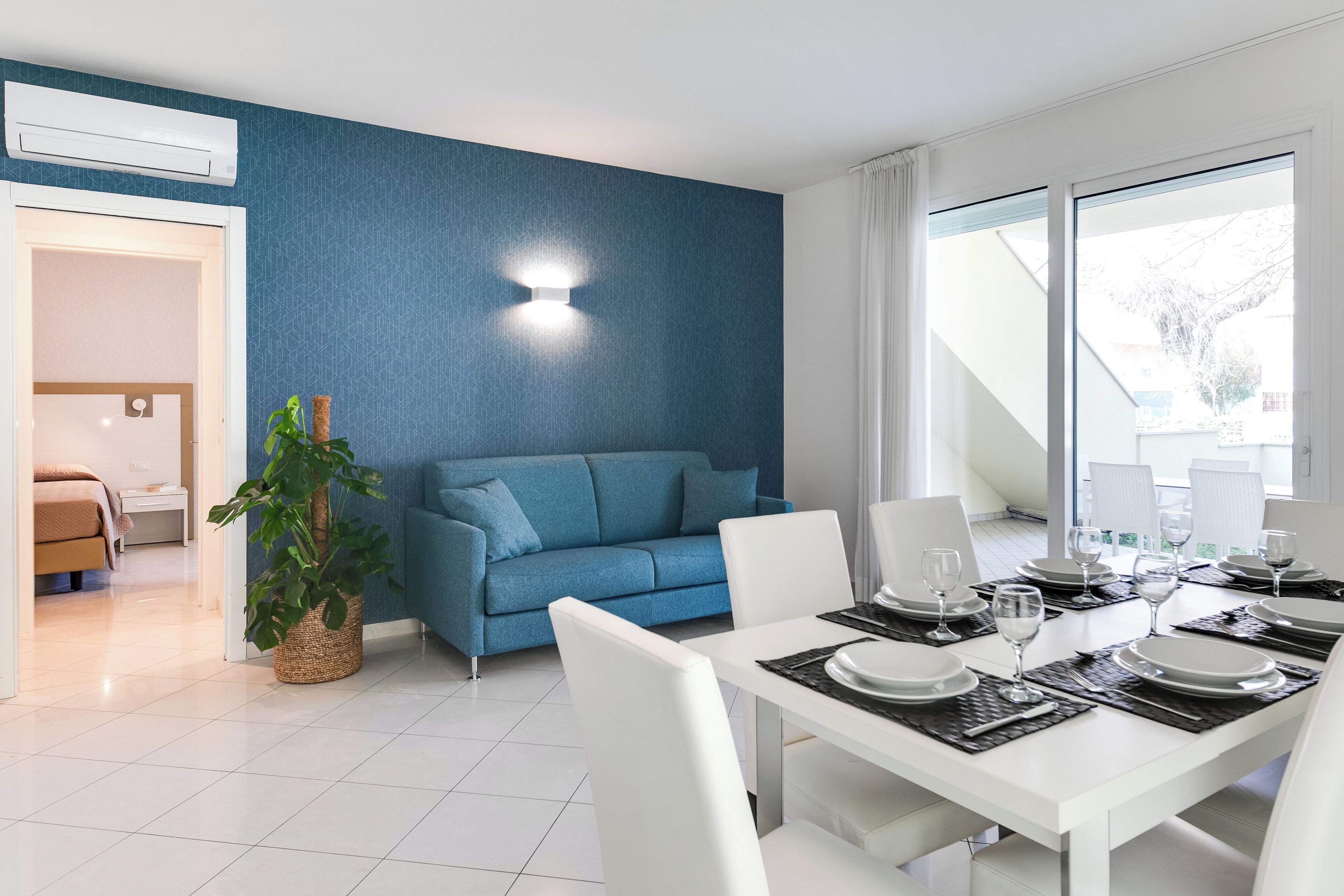 A modern dining area with a white table set for six, a blue sofa, and a large window offering a view of the outdoors.