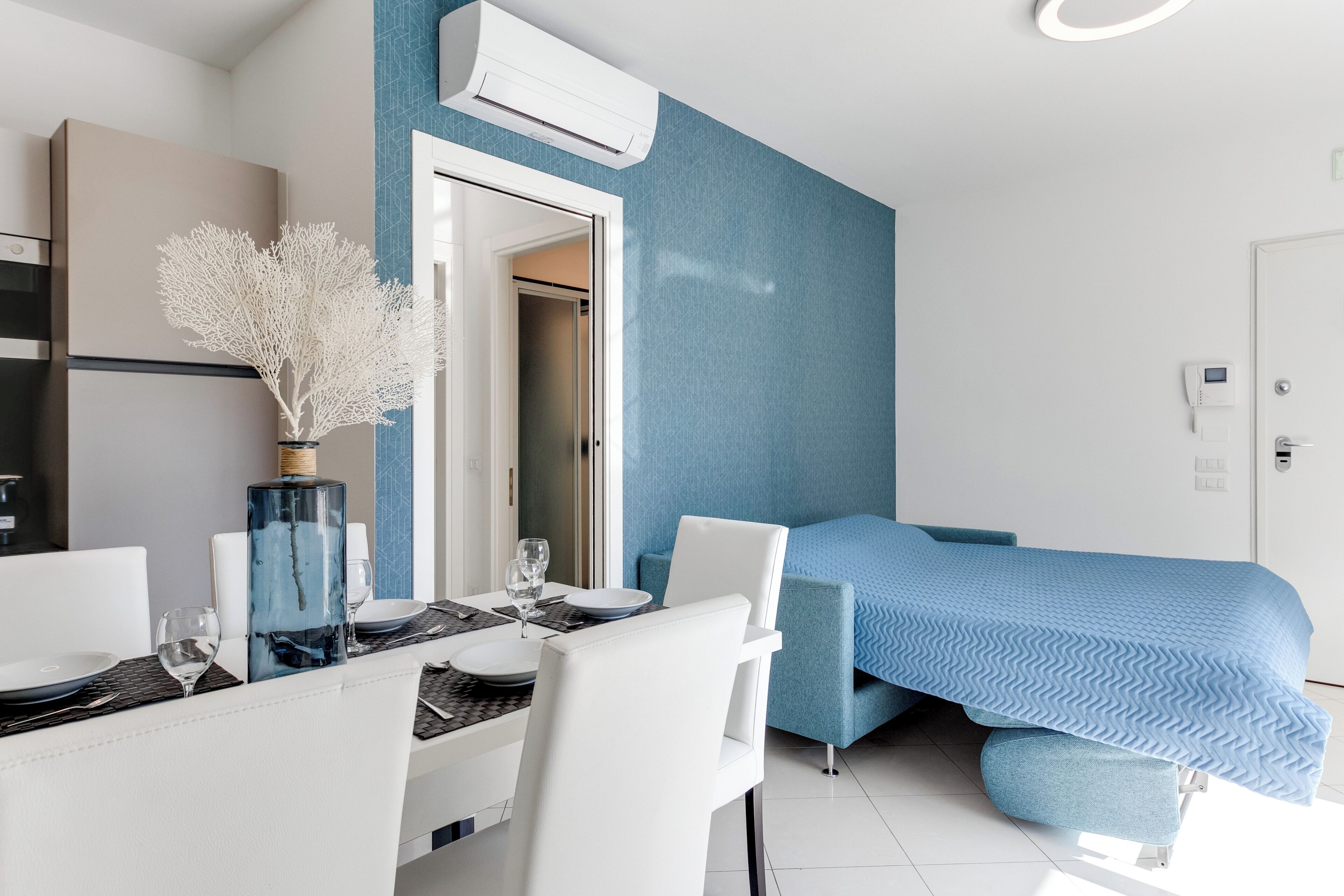 Una habitación de hotel moderna con comedor, una cama con colcha azul y un equipo de aire acondicionado en la pared.