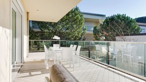 A balcony with a glass railing, white furniture, and a view of neighboring buildings and trees. - Residenza Acquamarina - Siroli Collection (Cervia)