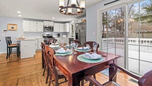 Un coin repas avec une table en bois dressée pour six personnes, des armoires blanches, un réfrigérateur en acier inoxydable et une grande fenêtre donnant sur une courClose.