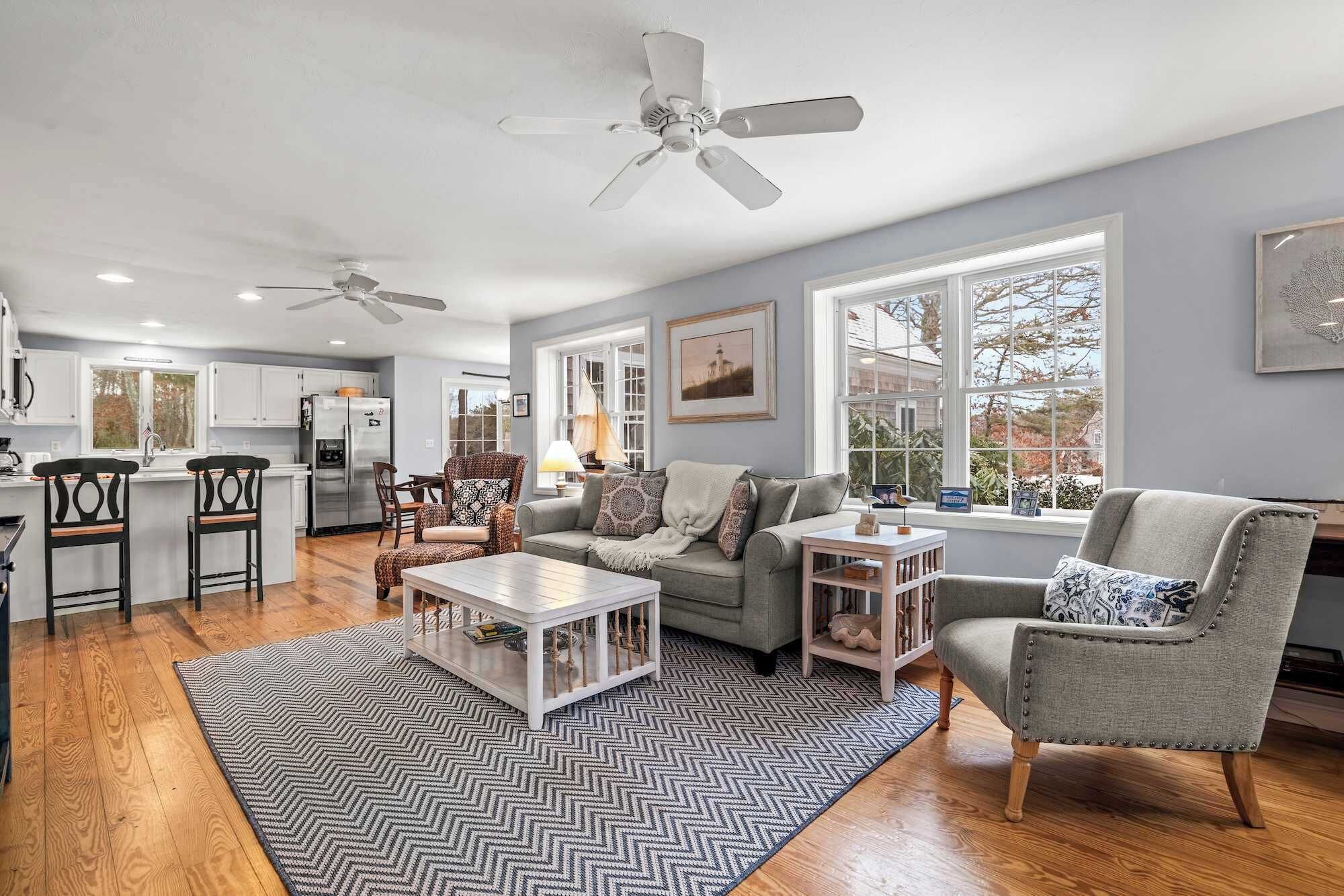 A spacious living area with a grey sofa, a coffee table, and a dining table with chairs. There are ceiling fans, a kitchen in the background, and large windows with a view.