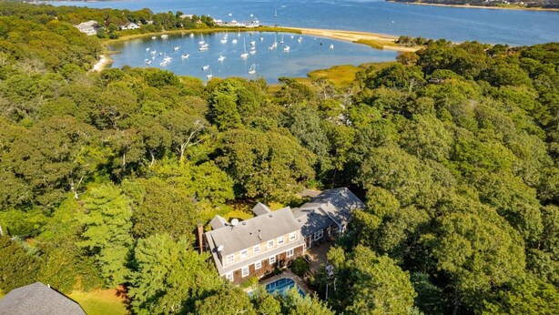 Une grande maison entourée d’arbres denses, un plan d’eau avec des bateaux et une plage en arrière-plan.