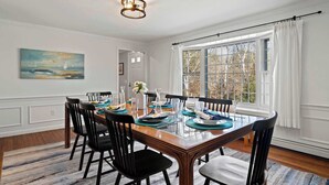 Une salle à manger avec une table en bois dressée pour six personnes, des chaises noires, un tableau au mur et une grande fenêtre avec des rideaux.