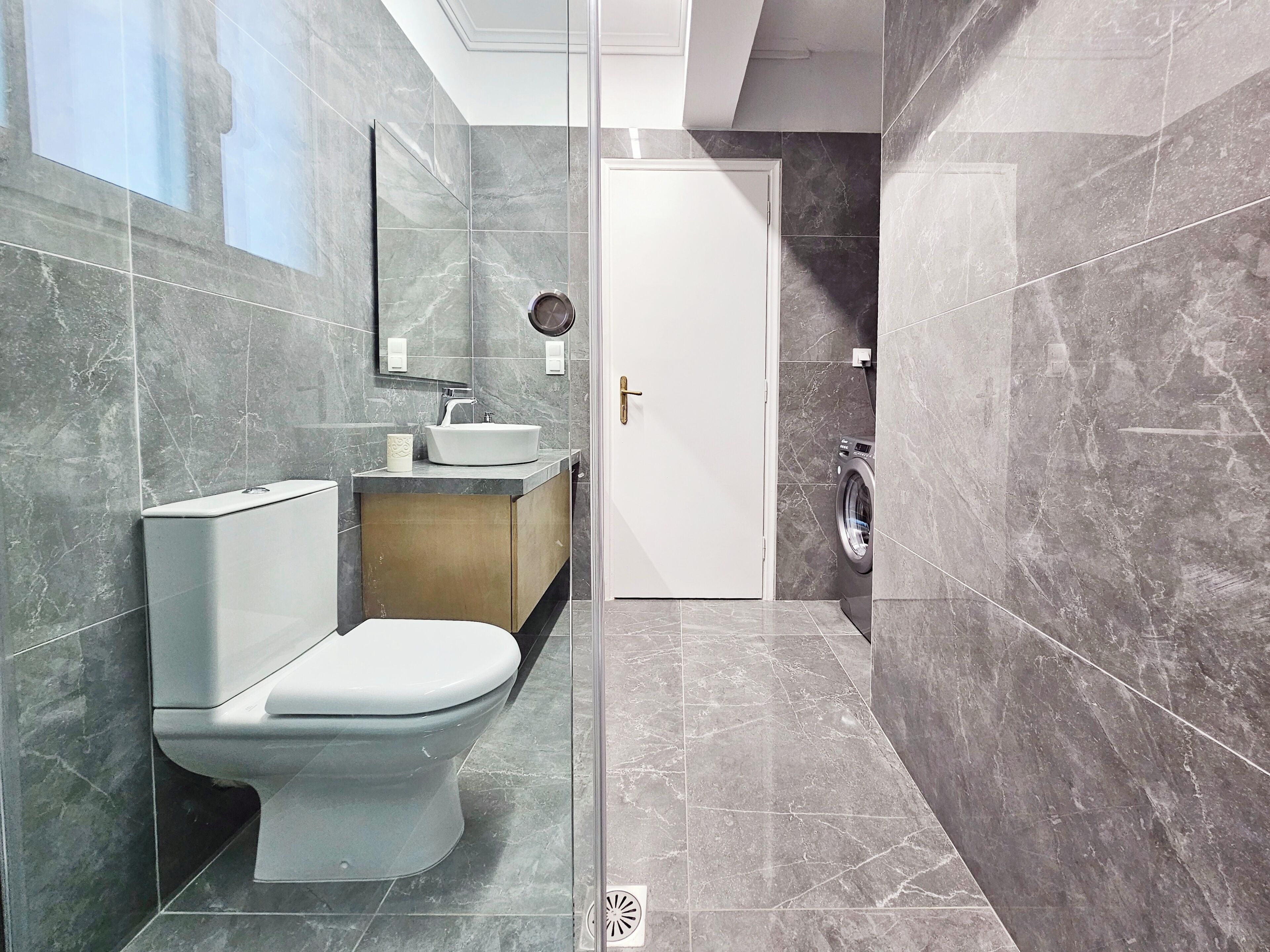 Une salle de bain moderne équipée de toilettes, d’un lavabo et d’un lave-linge, avec des murs en marbre et une cabine de douche en verre.