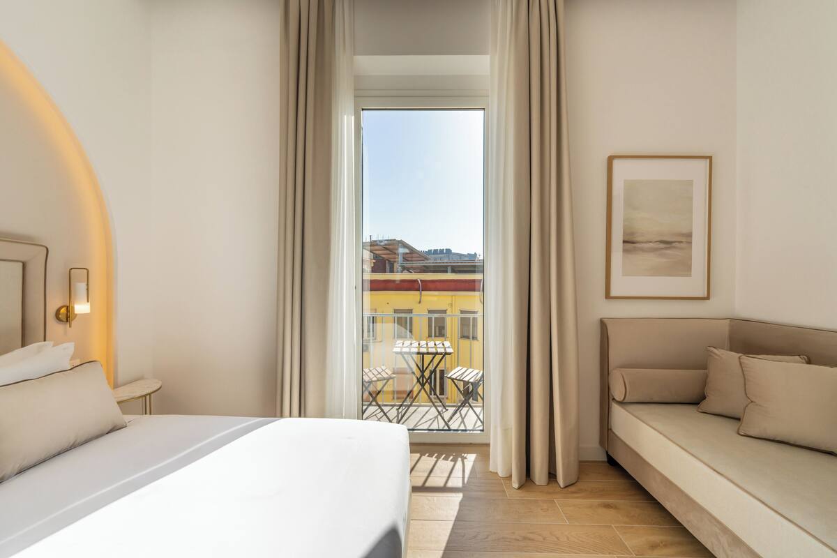 A hotel room with a bed, a sofa, a balcony with a table and chairs, and a framed picture on the wall.