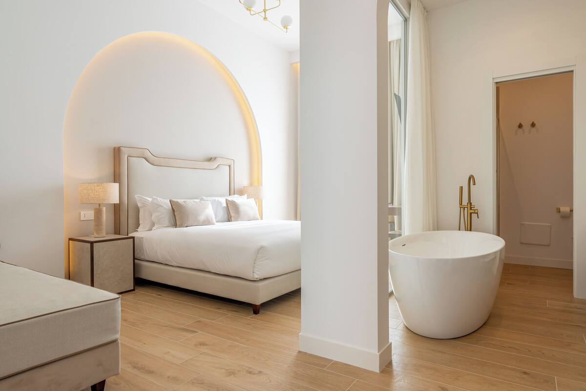 A modern bedroom with a large bed, bedside tables, a large white bathtub, and a view of the bathroom.