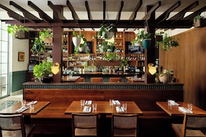 Un restaurant avec des tables et des chaises en bois, un bar avec des étagères remplies de bouteilles et des plantes suspendues.