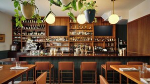Un área de bar con estantes de madera llenos de botellas, una barra equipada con utensilios y plantas colgantes. Mesas y sillas dispuestas para sentarse.