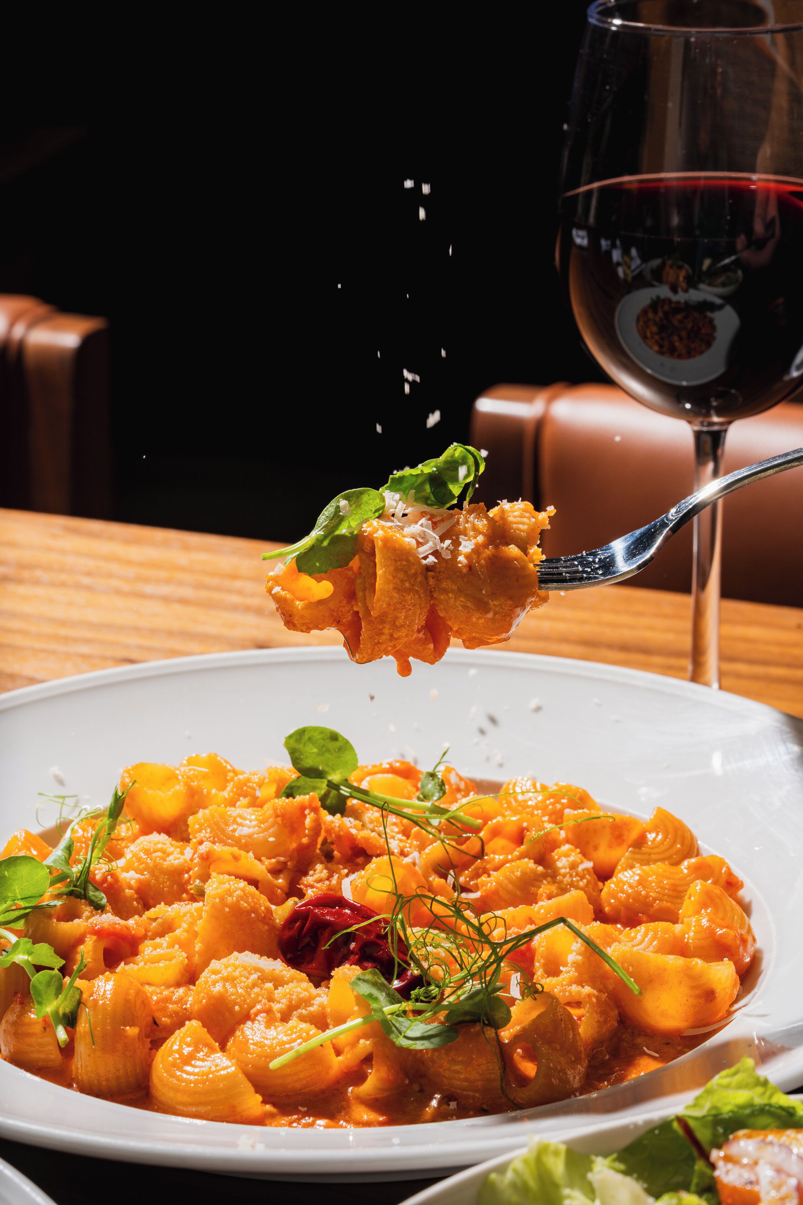 A plate of pasta with a fork lifting a piece, garnished with greens, and a glass of red wine in the background.