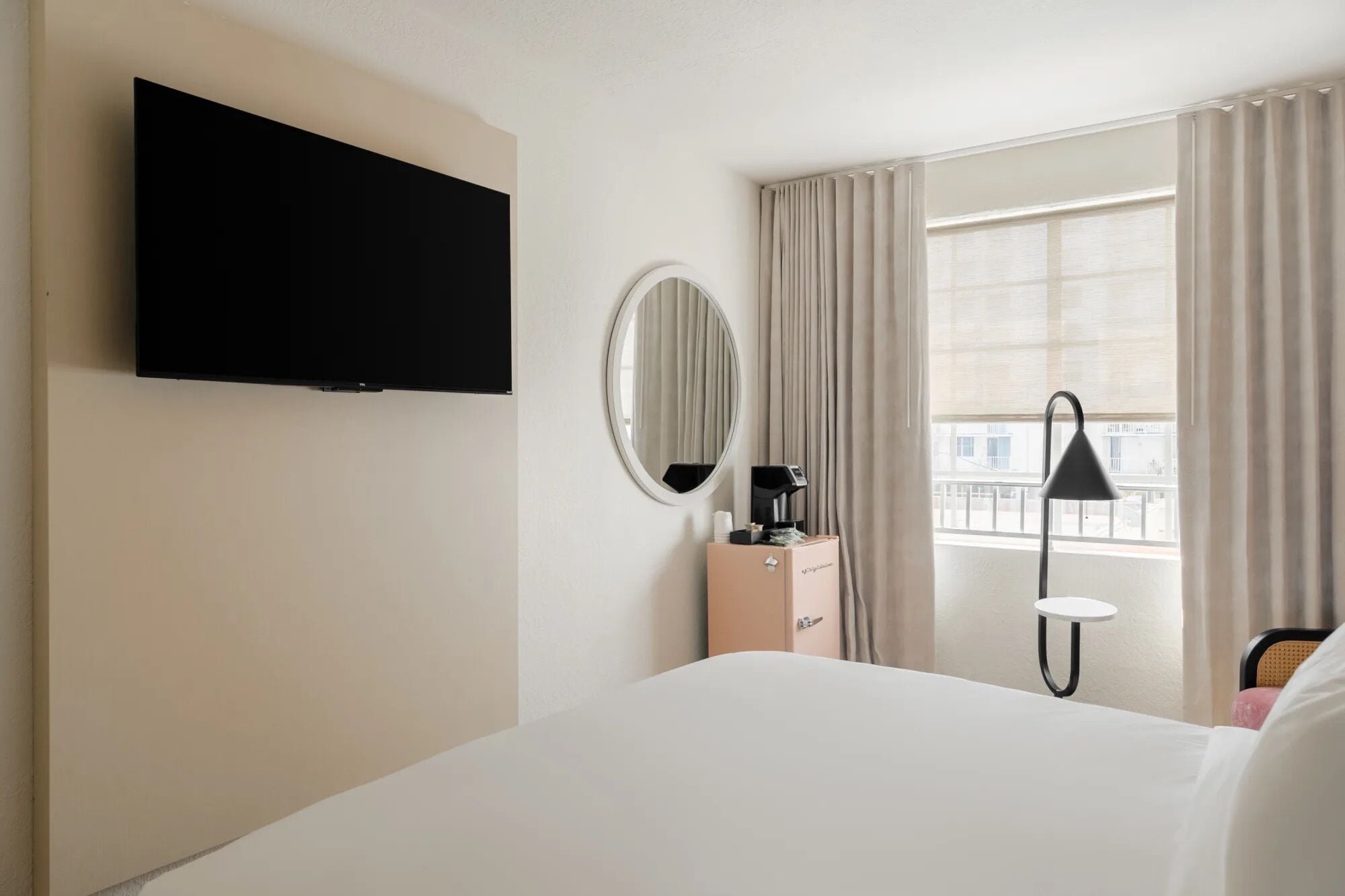 A hotel room with a bed, a TV mounted on the wall, a bedside table with a coffee maker, a lamp, and a window with curtains.