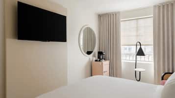 A hotel room with a bed, a TV mounted on the wall, a bedside table with a coffee maker, a lamp, and a window with curtains.