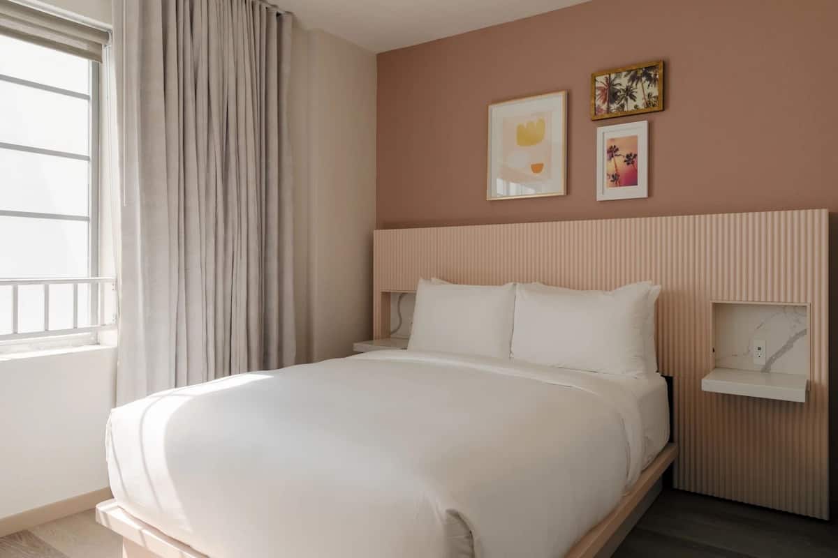 Camera d'albergo con un letto grande, un comodino, una mensola a muro e una finestra con tende.