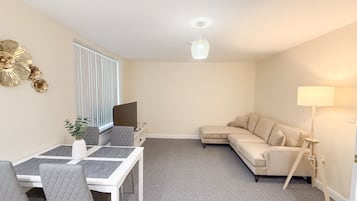 A modern living room with a dining area, a beige sofa, a standing lamp, and a TV.