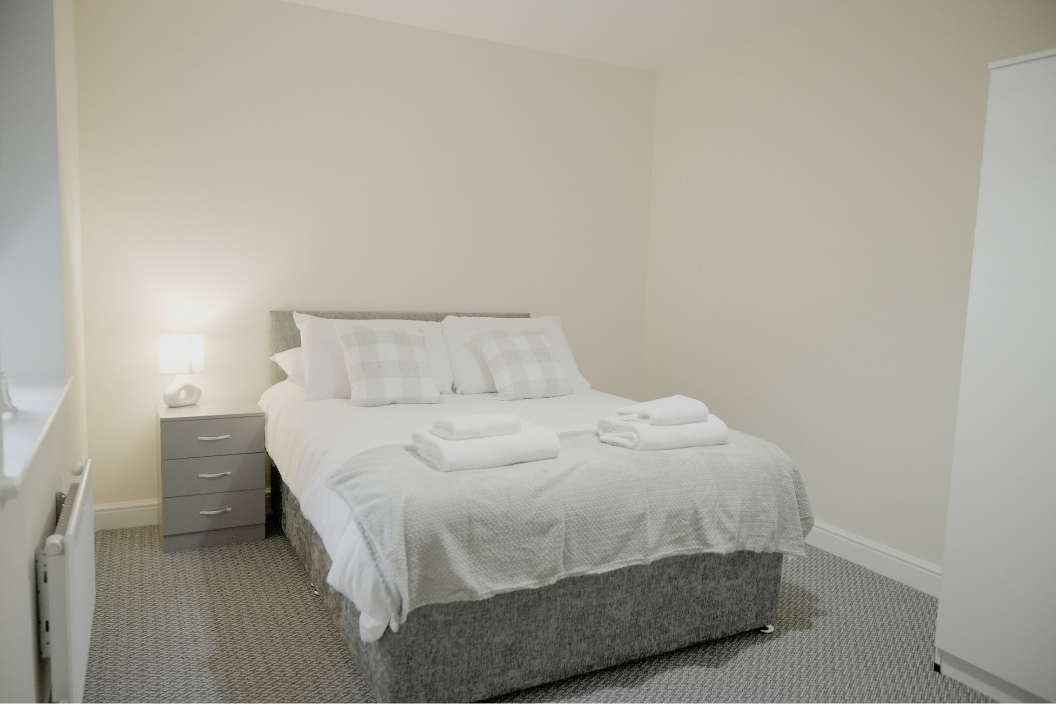 Un lit bien fait, avec du linge de lit blanc et une tête de lit grise, une table de chevet avec une lampe, et un mur blanc sobre.