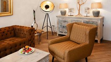 A living room with a brown sofa, a quilted armchair, a dining table with a fruit plate, a floor lamp, and a decorative cabinet with lamps.