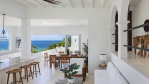 A spacious living area with a large window overlooking the ocean, a dining area with wooden furniture, and a white sofa. - VILLA SANTORINI - walk to beach and restaurants from this luxurious masterpiece (Tortola)