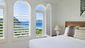 A bedroom with a large bed, a nightstand, and a lamp. There are three windows with shutters, and a view of the ocean and greenery. - VILLA SANTORINI - walk to beach and restaurants from this luxurious masterpiece (Tortola)