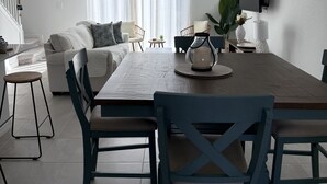 A modern living room with a dining area, featuring a wooden dining table, blue chairs, a sofa, and a TV mounted on the wall.