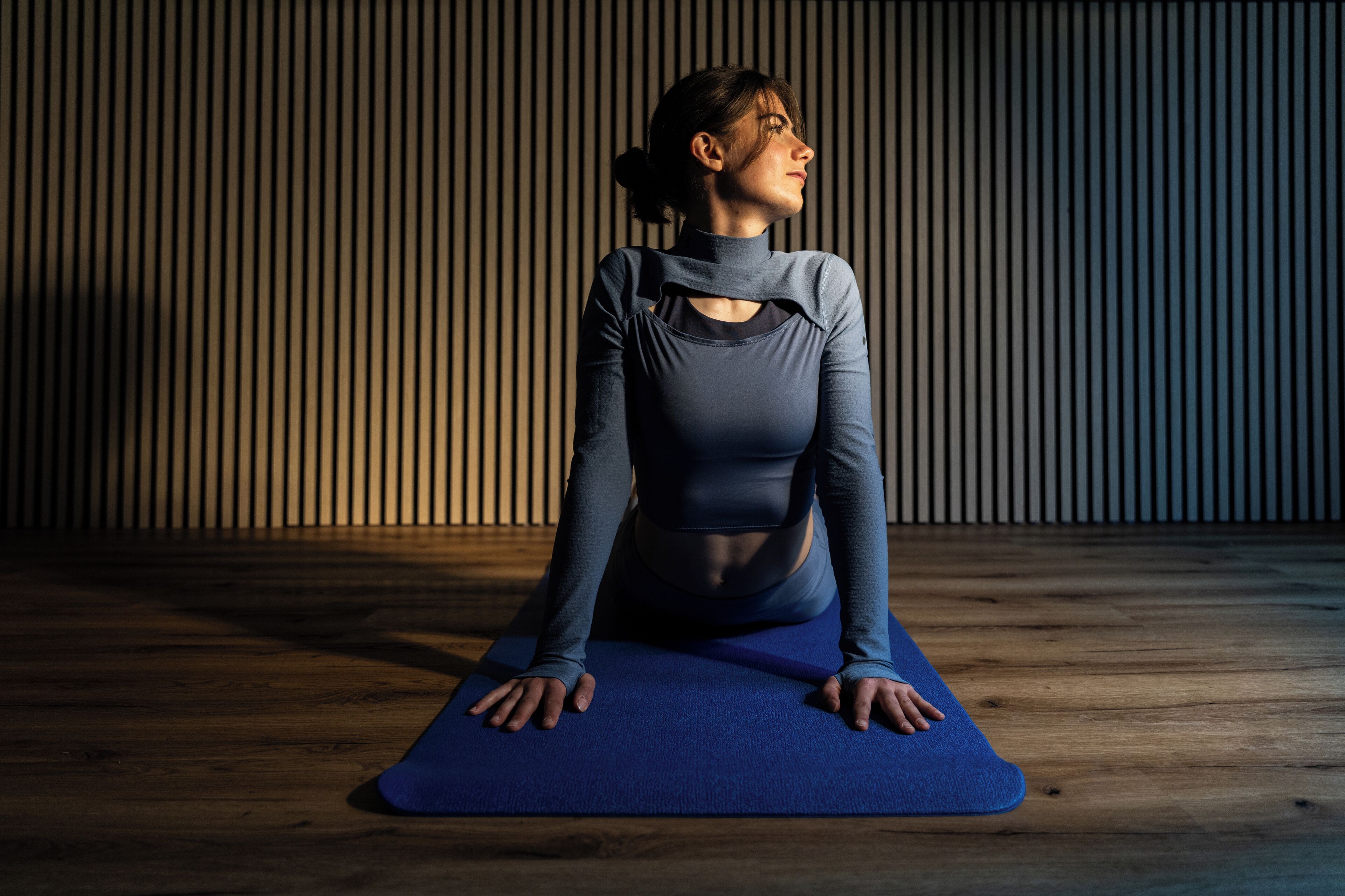 Uma pessoa sentada num tapete de yoga azul numa divisão com soalho de madeira e persianas de lamelas verticais.
