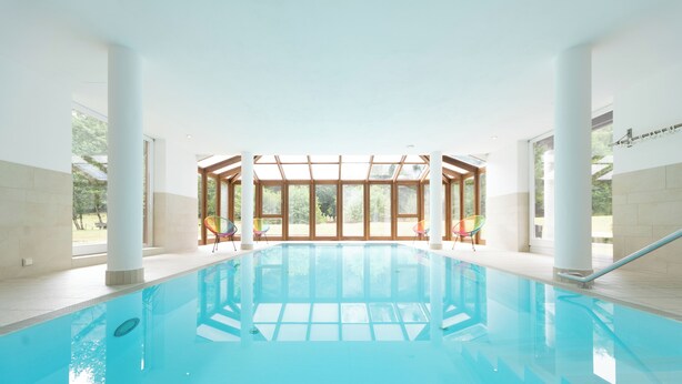 Una piscina interior con paredes de vidrio transparente, suelo de baldosas y vistas a zonas verdes.