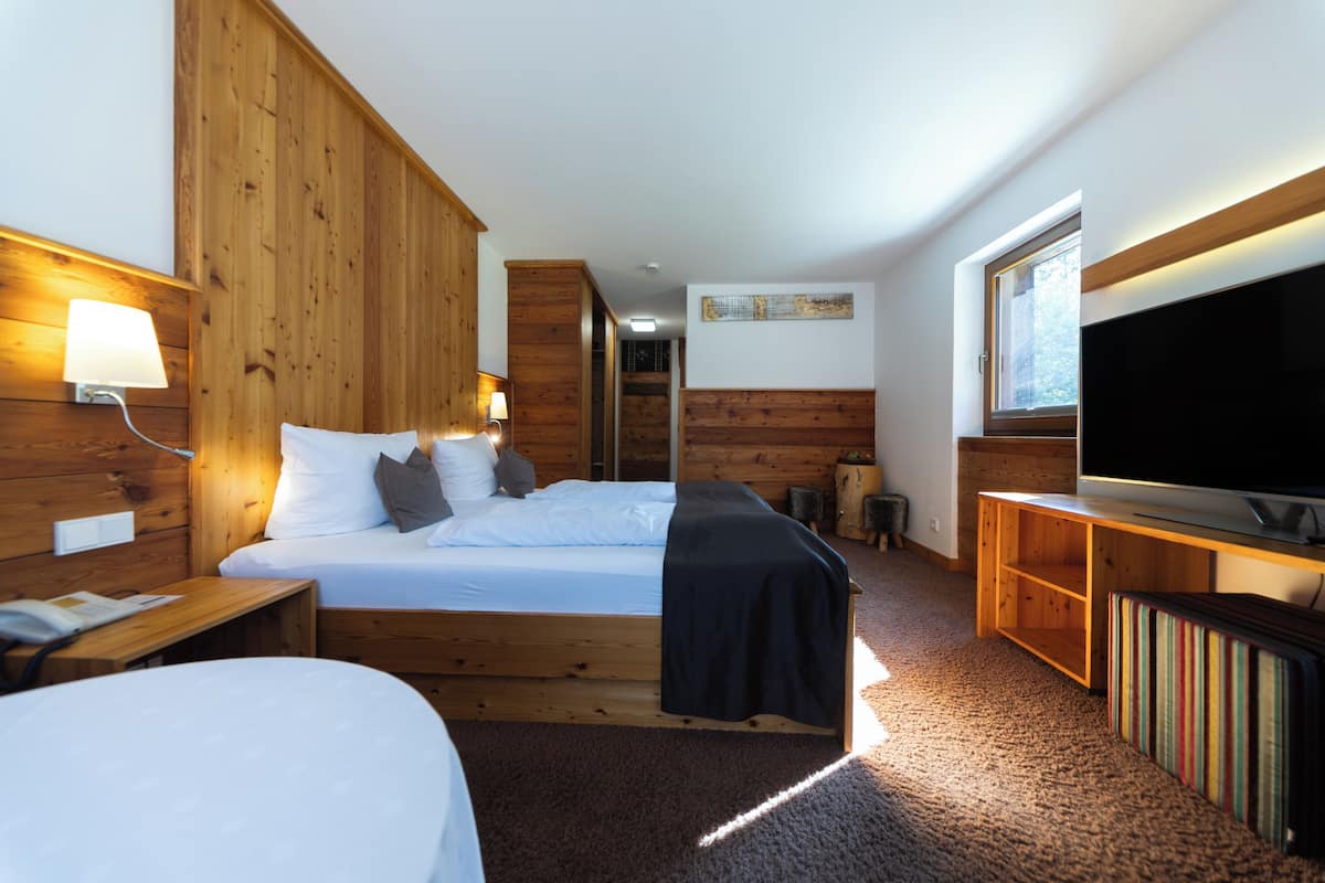 A bedroom with a wooden bed, a bedside table, a lamp, a TV, and a window.