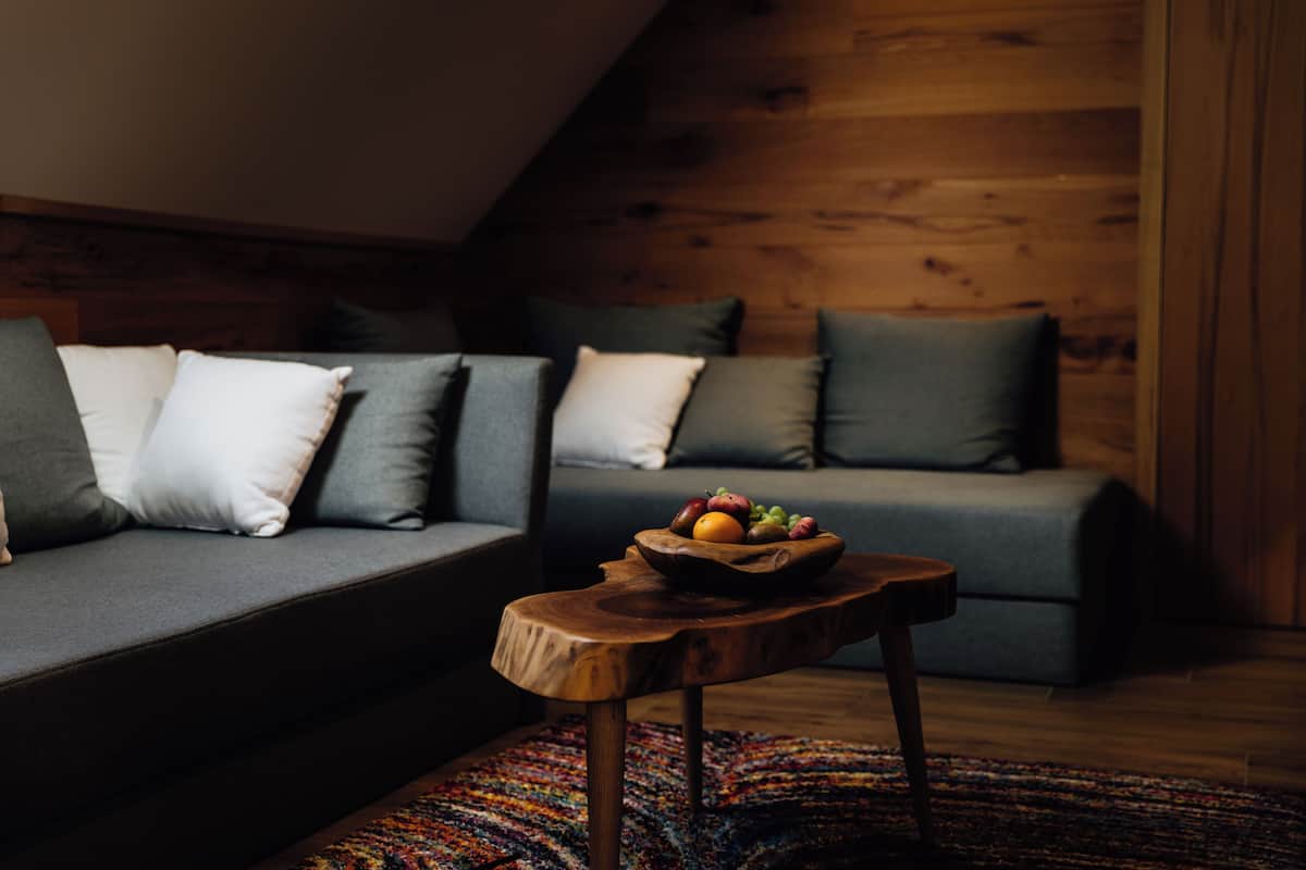 A cozy living room with a wooden ceiling, a grey sofa with cushions, and a small wooden table with a fruit bowl.