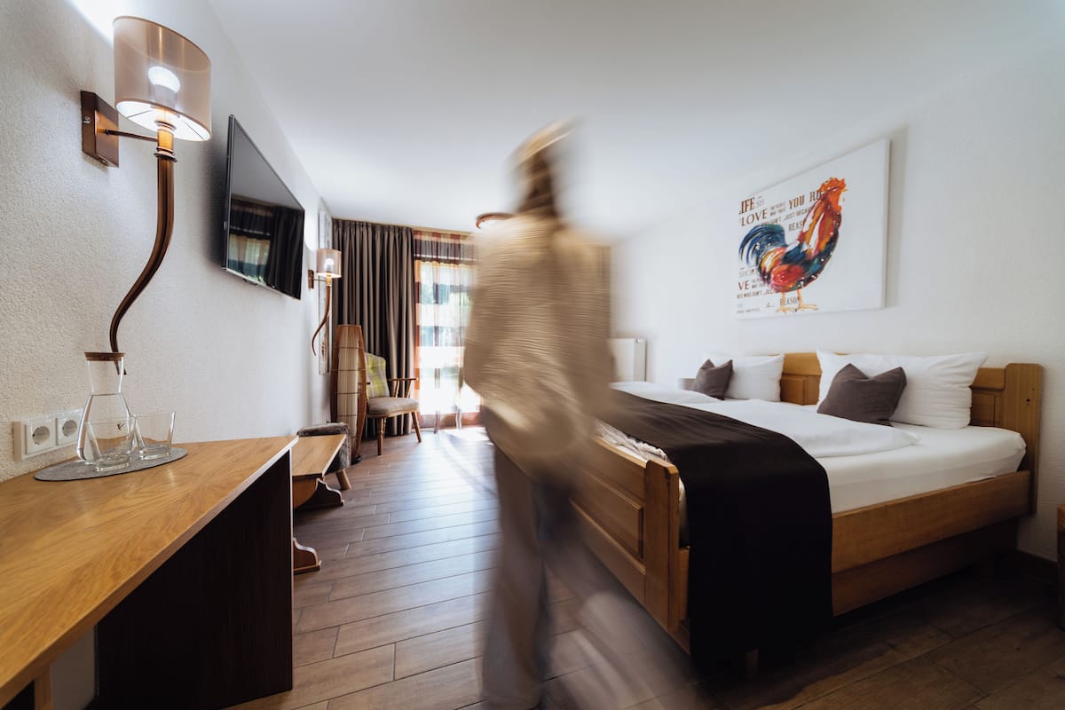 A hotel room with a bed, a desk with a lamp, a chair, a window with curtains, and a wall-mounted TV.