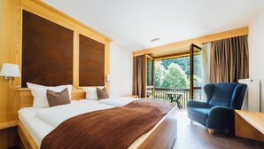 Habitación de hotel con una cama grande, un cabecero de madera, un sillón azul y un balcón con vistas a árboles.
