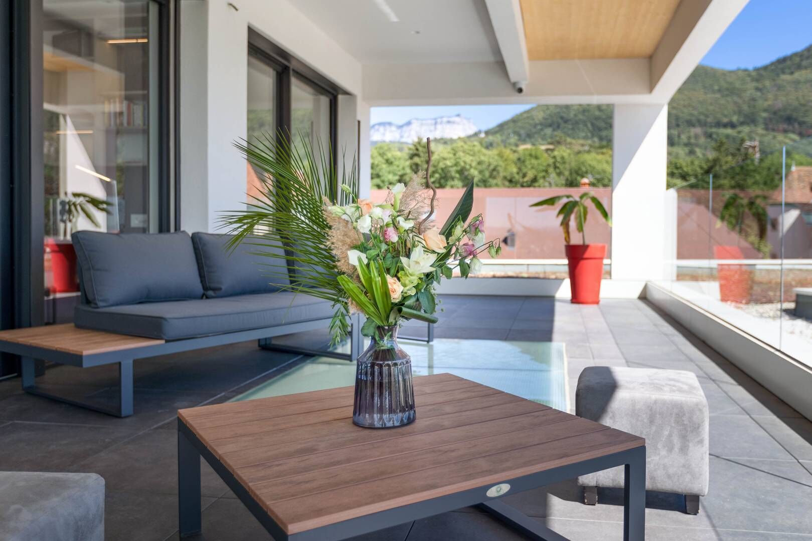 A modern outdoor seating area with a sofa and a table, a vase with flowers, and a view of mountains.